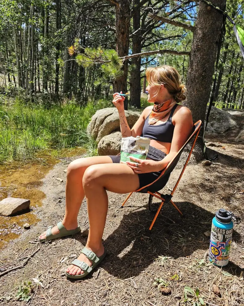 A camper in a camp chair by a creek eating Oso Posole from the pouch