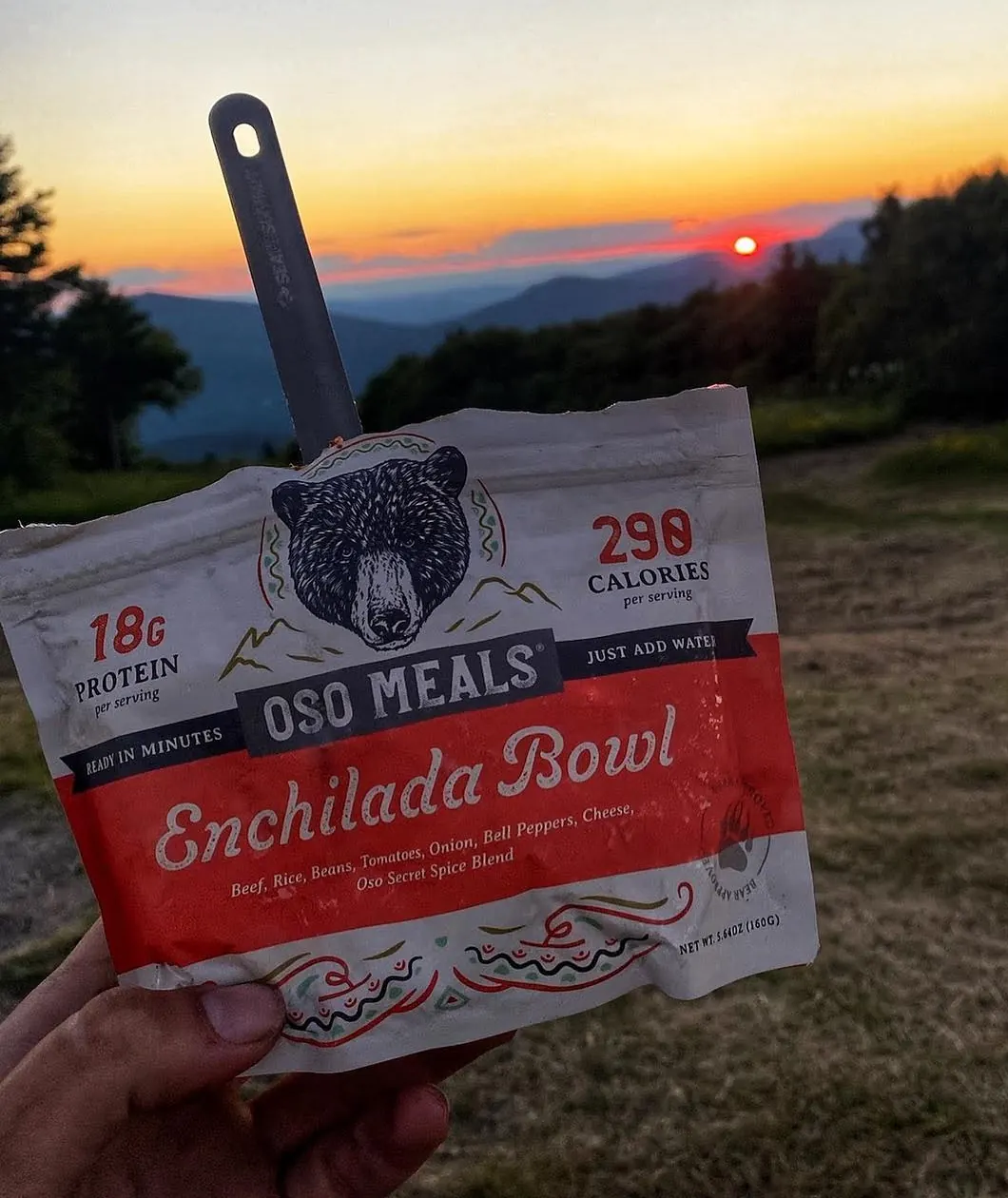An Oso pouch with a spoon in it, held up at sunset over the mountains