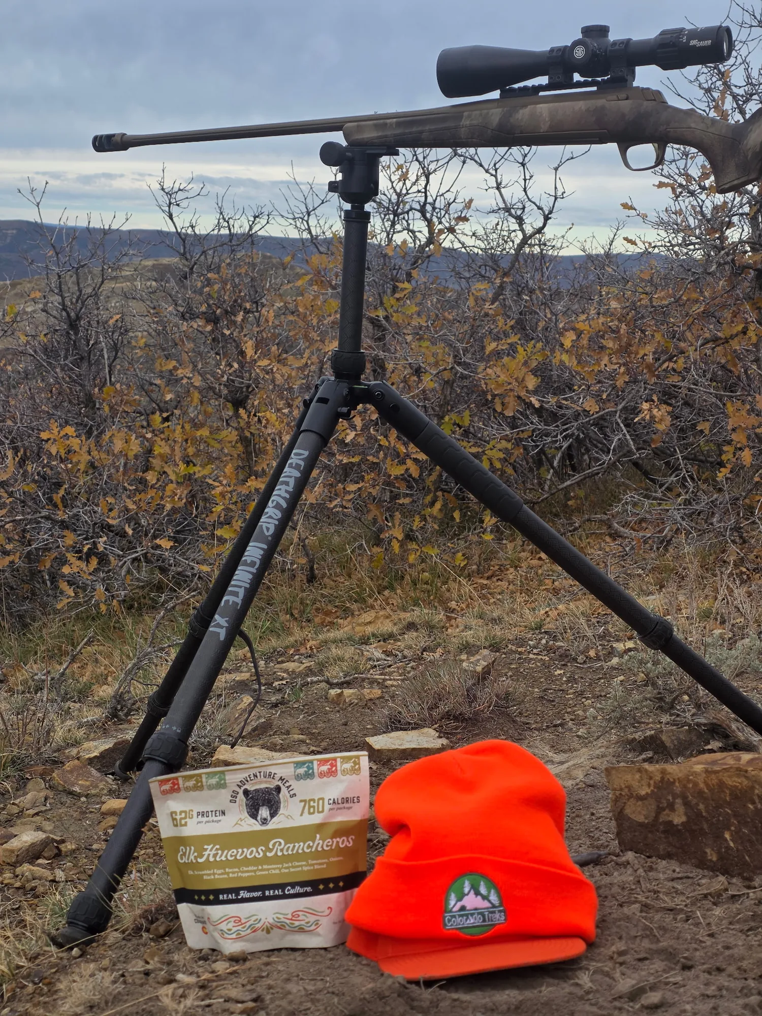 An Oso Elk Huevos Rancheros pouch and a blaze orange beanie at the foot of a rifle on a shooting tripod, with oak brush and mesas behind