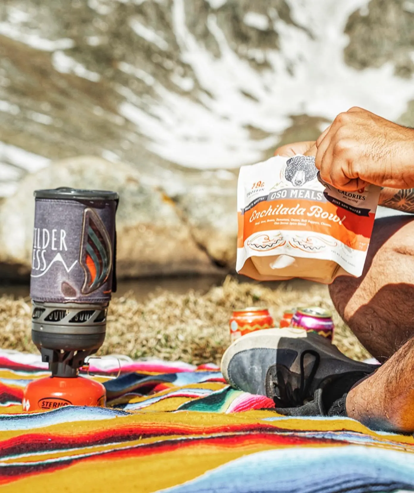 A backpacking stove heating water on a serape blanket while someone opens an Oso pouch